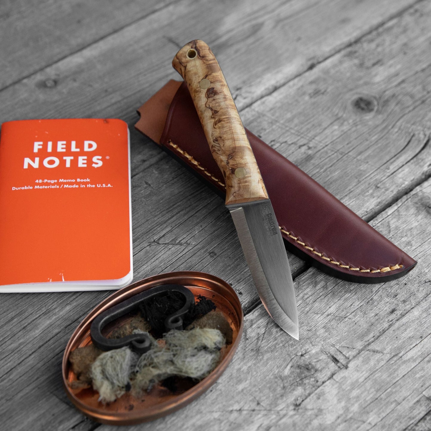 Casström Woodsman knife in curly birch, resting on a leather sheath and with other outdoor equipment in the image (notebook, flint striker, tin).