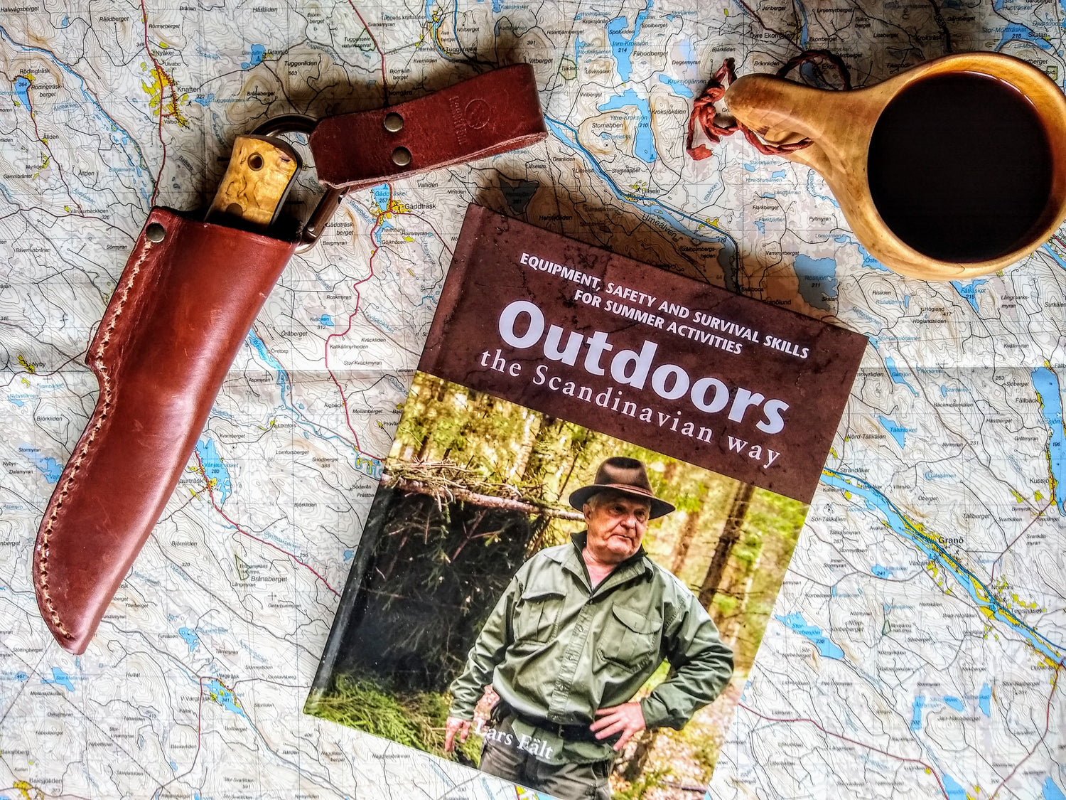 Book titled 'Outdoors the Scandinavian way' on a map with a knife sheath and kuksa cup of coffee.