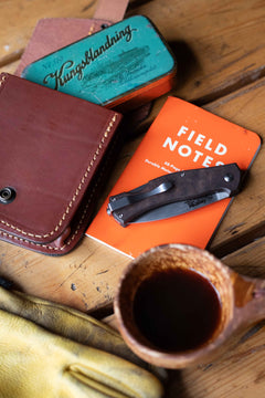 Casström Lars Fält EDC folding knife resting on a Field Notes notebook with leather possibles pouch, tobacco tin, and kuksa cup of coffee.