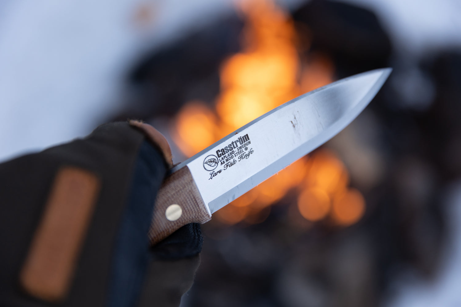 A gloved hand holding the Casström Lars Fält knife with canvas micarta handle, with a campfire in the background.